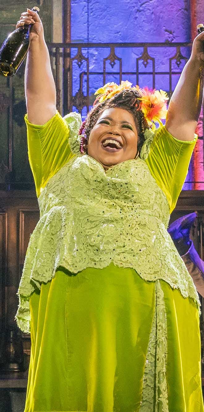 Persephone stands in a bright green dress; her arms are raised and a bottle of wine is in each hand.