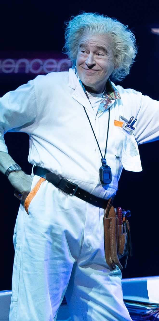 Don Stephenson as Doc Brown stands with hand on hip and smirk on his face.