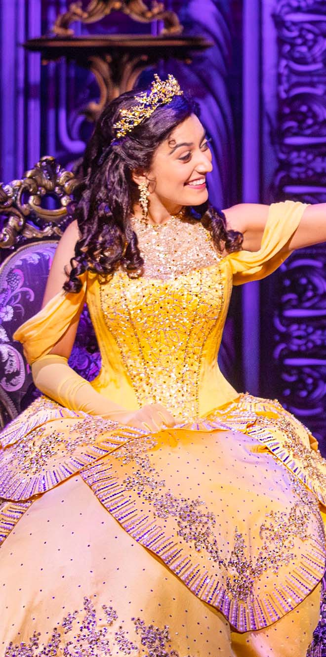 Bell sits in an ornate chair while wearing her icon yellow ball gown, smiling and looking to the right.