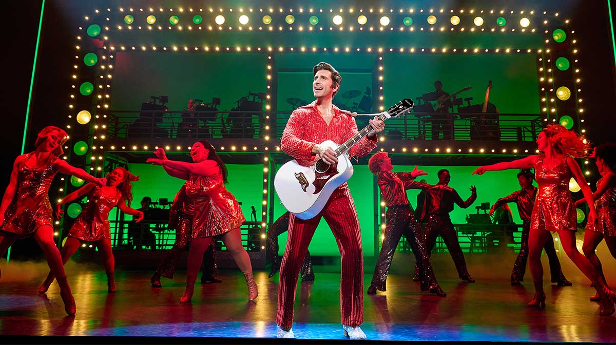 Nick Fradiani as Neil Diamond holds a white acoustic guitar while wearing a red sequin outfit. Dancers appear on stage behind him.